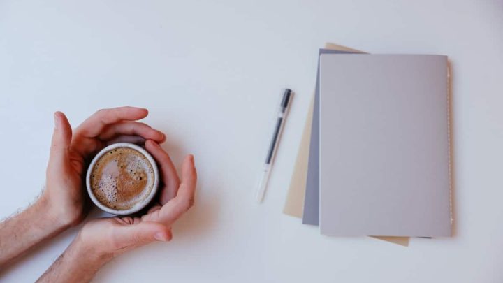 A man wraps his palms around a cup of coffee with a few papers and a pen kept at a distance from him