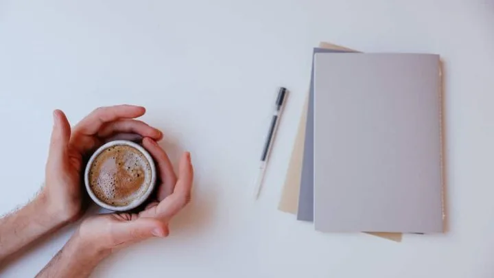 A man wraps his palms around a cup of coffee with a few papers and a pen kept at a distance from him