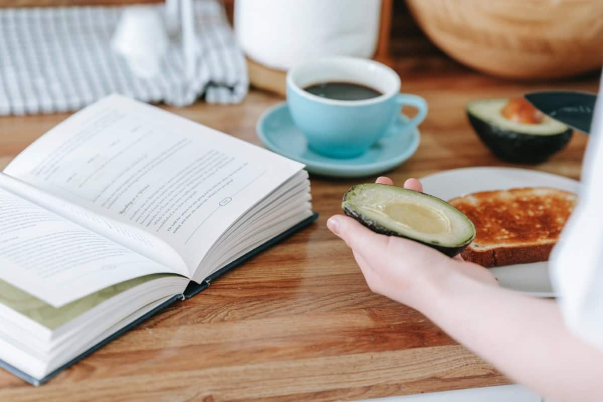 A woman with a avocado in her hand in front of a breakfast table with coffee, toast and a book. 