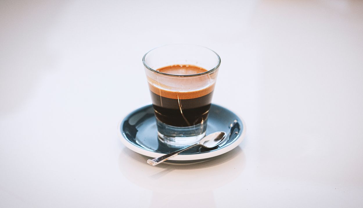 An espresso shot in a clear glass atop a saucer with a teaspoon.