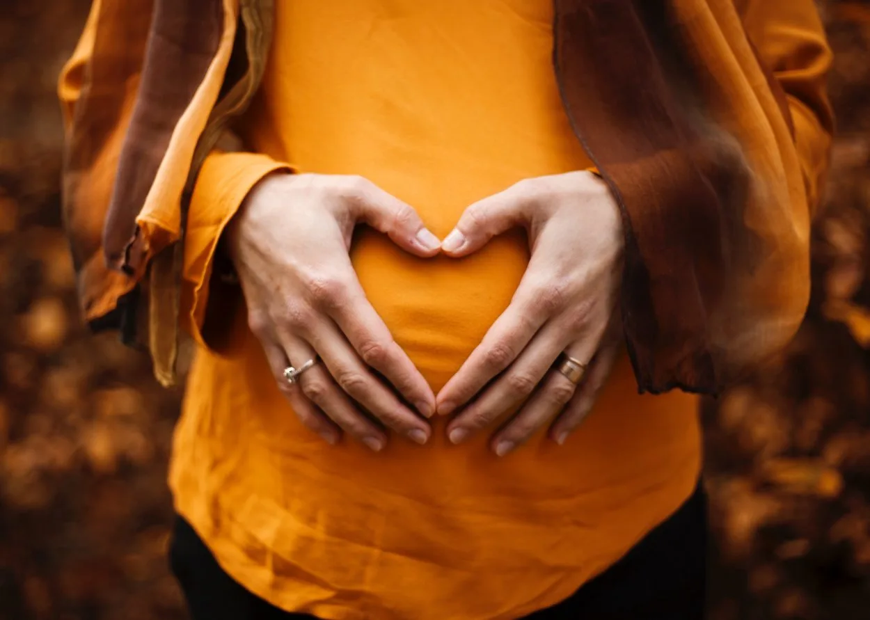 a women pregnant standing in the fall leaves with a hand heart on her belly