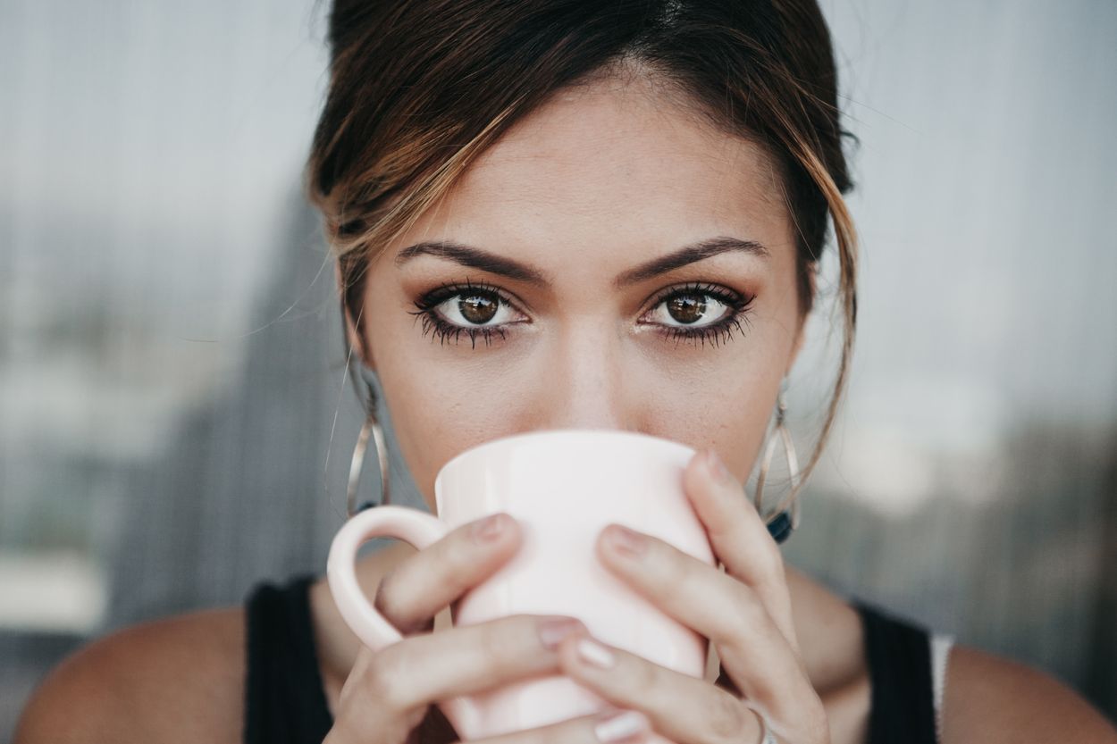 A woman looking at the camera while holding a white mug of coffee and drinking it.