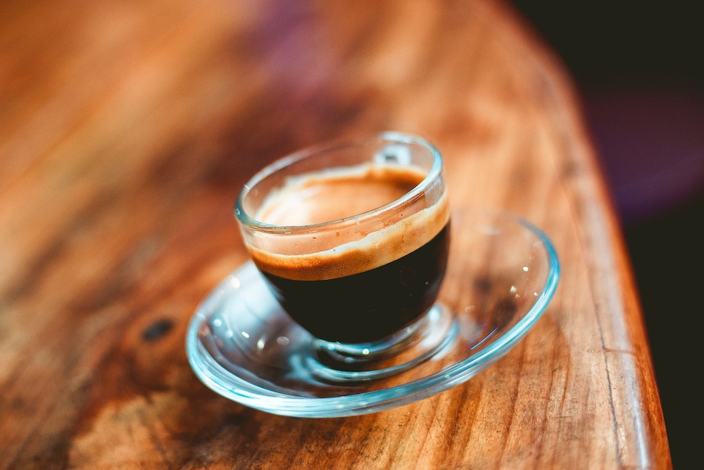 A freshly pulled espresso with beautiful crema in a clear glass atop a clear saucer.