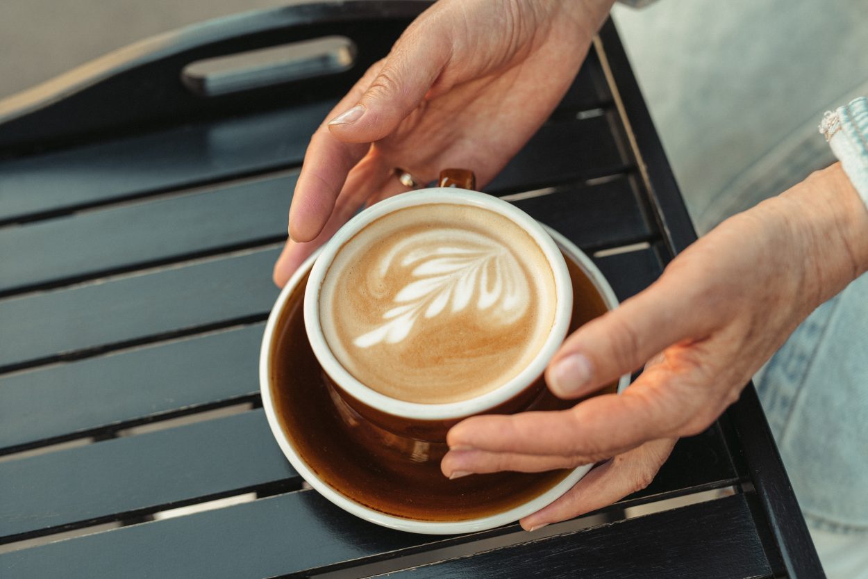 A cup of red cappuccino with latte art in a brown cup and saucer being held by a woman.