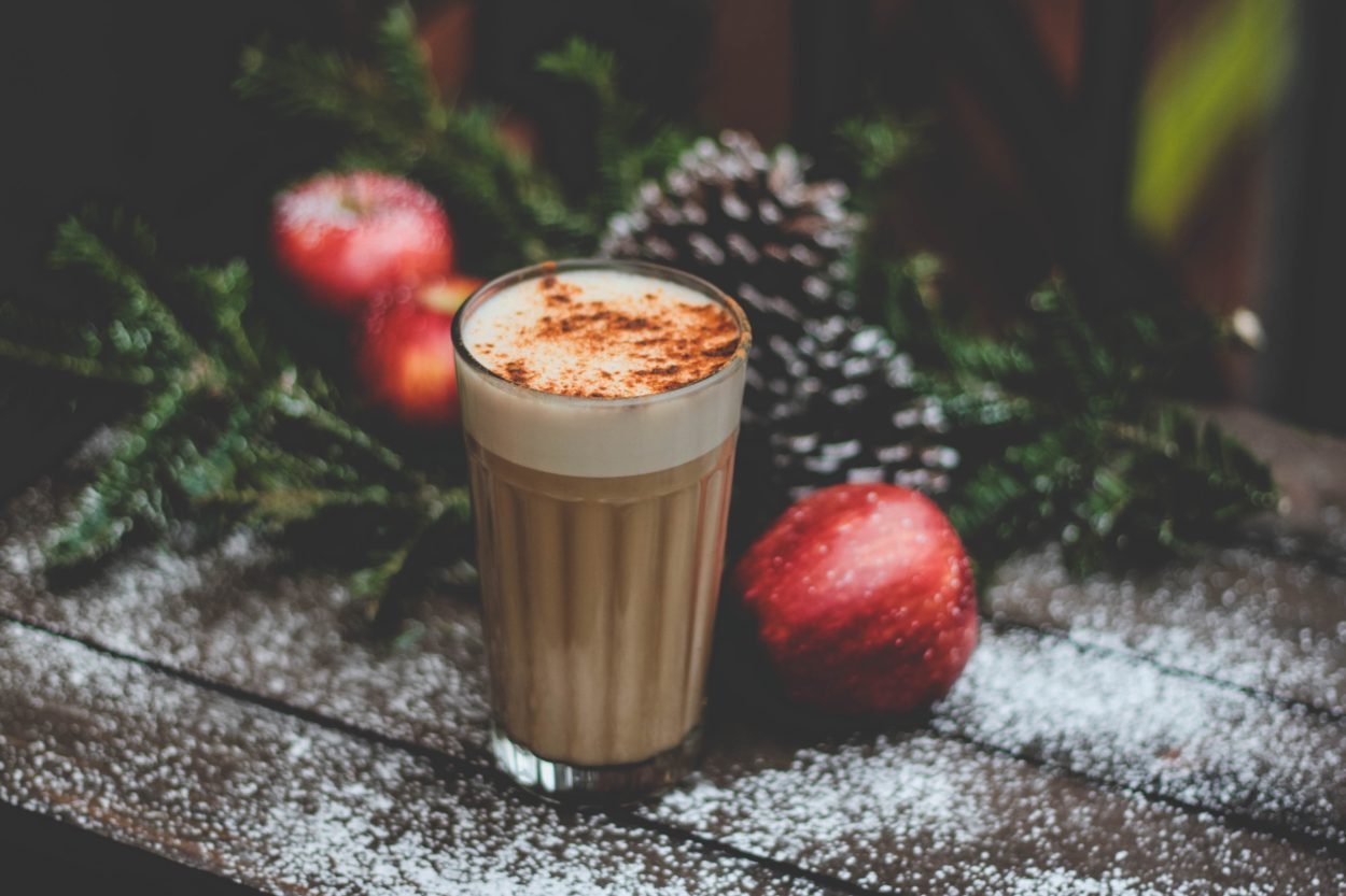 A cappuccino with chocolate powder sprinkled on top, with apples and pine leaves on the background.