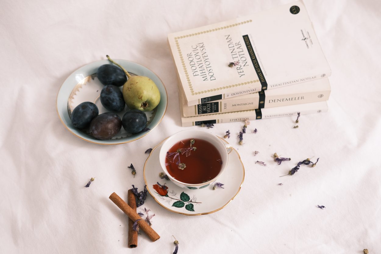 A herbal tea or tisane in a white cup atop a saucer with floral design with books and fruits beside it.