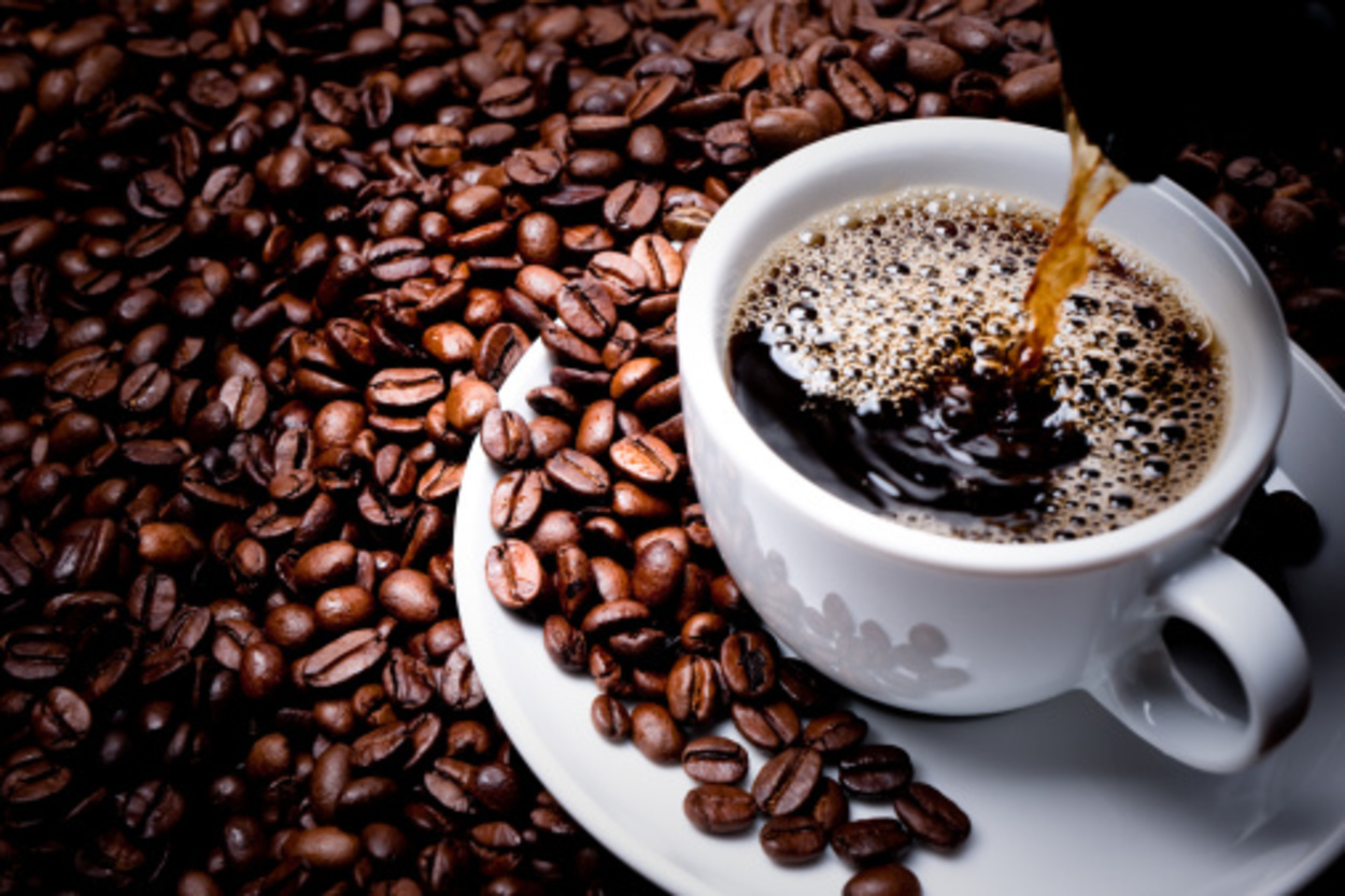 Lots of coffee beans are scattered around and black coffee is being poured in a white cup.