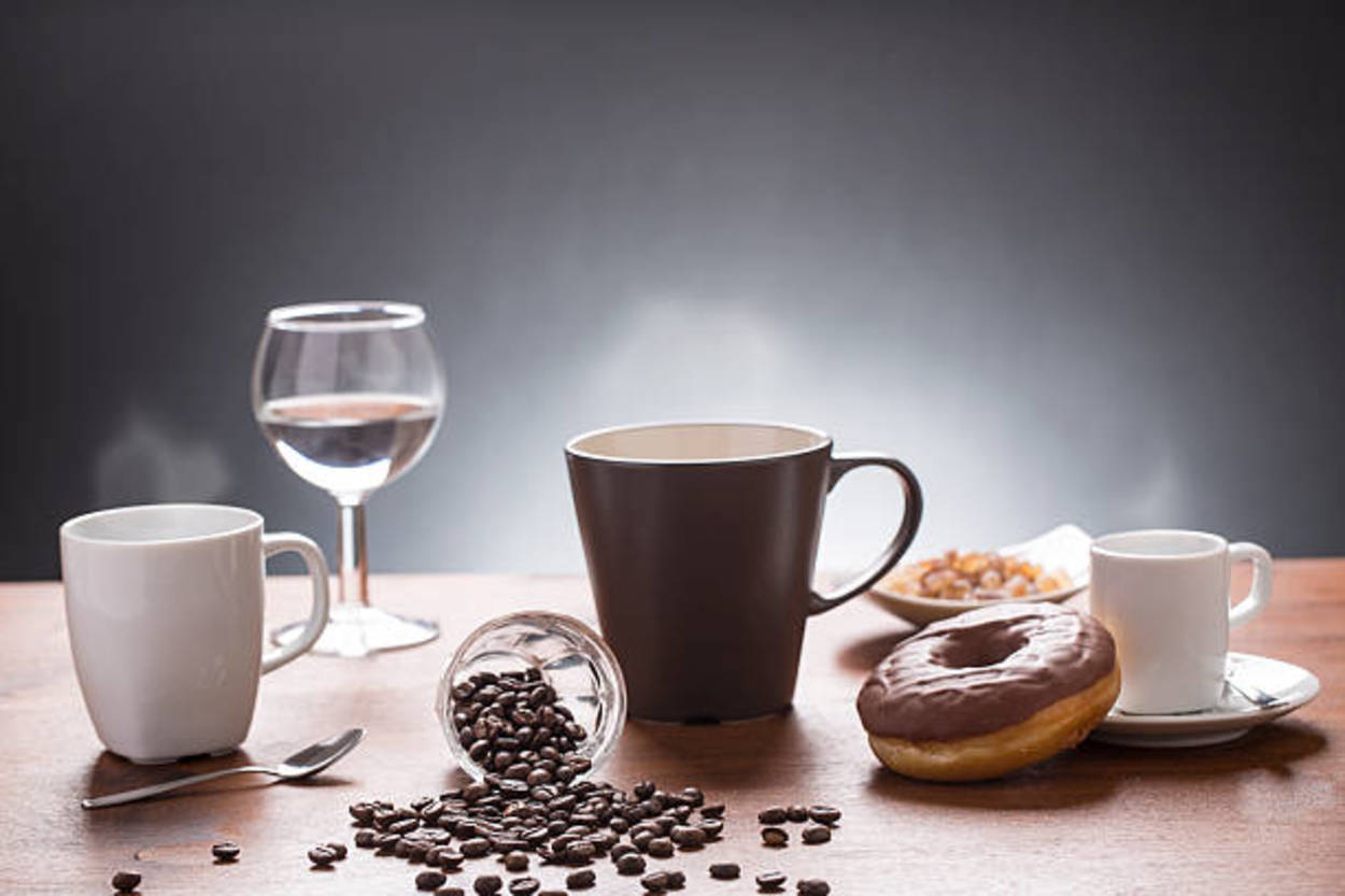 A wooden table with many things on top like a spoon, a donut, a wine glass half-filled with water, a white mug, a brown mug, and a small container containing coffee beans that have spilled over.