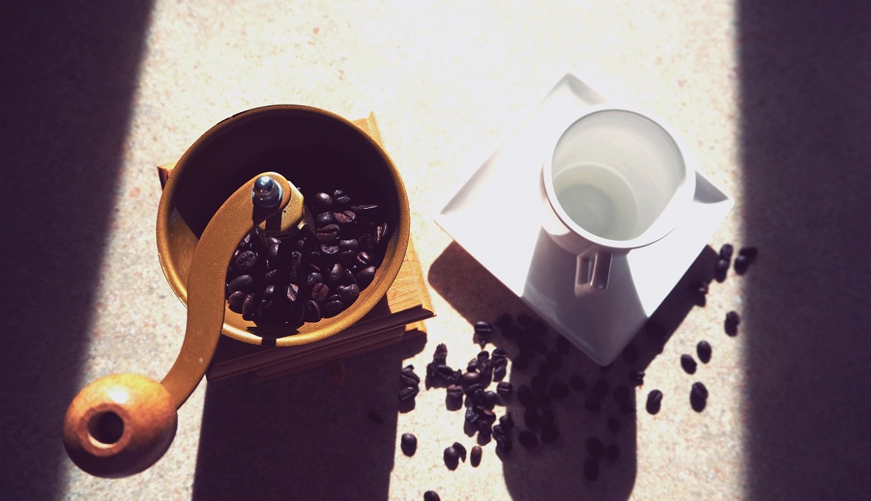 An image of coffee beans in a manual coffee grinder and a cup placed near by.